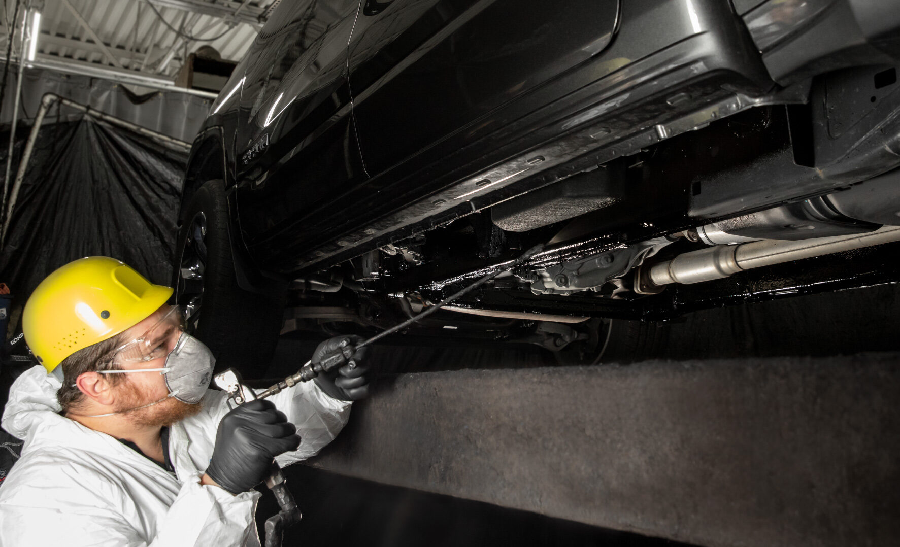 undercoating_process_2_bottom A worker in a black shirt and gloves inspects a blue car's rear bumper closely in a well-lit garage environment